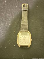 Photo of a vintage Timex wristwatch with a gold-tone square case, white dial featuring analog hands and a top digital window, attached to a textured black leather strap with gold-tone buckle.