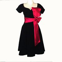 Front view of a 1950s black velvet party dress with a bright red satin sash tied in a large bow on the left waist, displayed on a mannequin.