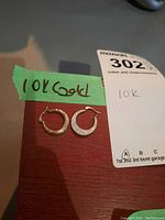 Pair of 10K gold earrings, one textured and one smooth, placed on a wooden surface with a green label reading '10K Gold'.