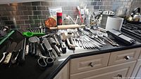 Wide view of kitchen counter showing an assortment of metal cutlery, kitchen utensils including scissors, peelers, graters, and a rolling pin arranged neatly.