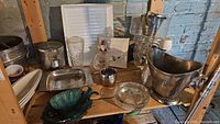 Wide view of shelf with metal champagne bucket, glass pitcher, crystal ashtray, oil bottle, metal cup, glass tumbler, and glass serving bowl.