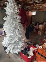 Large white artificial Christmas tree standing in basement area next to red bins filled with Christmas decorations.