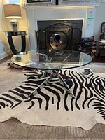 Glass top coffee table with a round clear glass surface and metal cross-leg base placed on a zebra pattern rug in a living room setting.
