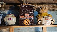Two President's Choice cookie jars and one vintage ceramic floral lidded jar shown on wooden shelf in basement setting
