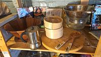 Wide shelf photo showing wooden salad bowl, stovetop coffee maker, bamboo steamer basket, wooden lazy Susan, wooden salad tongs, metal colander, glass parfait bowl, and four Ina Garten cookbooks.