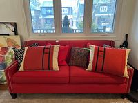 Red fabric couch with silver nail stud detailing, seven cushions of mixed patterns and sizes on couch, photographed from front with window behind.