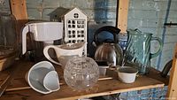 Wide shot of all items on wooden shelf showing kettle, ceramic teapot parts, crystal bowl, Brita pitcher, glass pitcher, and candleholder.