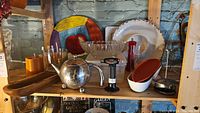 Photo shows shelf with atomic style metal teapot, pill mill, cooking torch, glass bowls, chip and dip set, and large ceramic plate with apple design in background