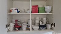 View of upper and lower shelves in cabinet containing various ramekins in red and green, white mugs and cups, small bowls, and glass vinegar bottle.