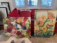 Two floral canvas prints displayed leaning against a red couch and nearby window light. One print shows multiple colorful flowers, the second has large orange and yellow flowers.