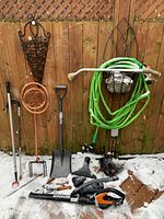 Full lot photo showing all gardening tools including the shovel, green hose on holder, Worx leaf blower, copper spiral plant support, hand cultivator, and decorative iron planter on wooden fence background with snow on ground.