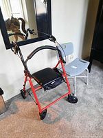 Red walker with black padded seat and support bar next to gray bath chair with backrest, positioned indoors on carpet.