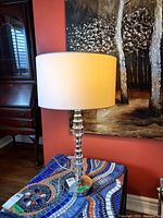 Glass table lamp with a stacked disc clear glass base and a white drum fabric shade placed on a colorful mosaic-tiled table, against a red wall with a painting in the background.