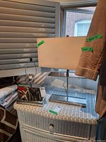 Two lamps placed on a light wicker-style cabinet. One lamp has a mirrored rectangular base and no shade, the other has a glass and metal base with a white rectangular fabric shade. Background includes window with blinds and hanging clothing.