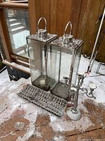 Two tall rectangular metal and glass lanterns with metal handles, a silver metal candelabra with five candle holders, and a rectangular plastic log holder on a wooden floor with snow