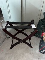 Wood luggage holder with foldable 'X' shaped legs and two black fabric straps across top, placed on concrete floor.