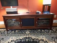 Front view of wood media unit with three doors: two glass panel doors and one central black mesh door. The unit has some scratches on the wooden surfaces.