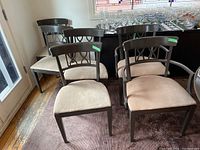 Six painted wooden dining chairs with upholstered beige seats and decorative backrests, two with arms and four side chairs, shown together on carpet near glass cabinet.