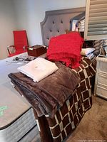 Photo showing stacked blankets and pillows on a bed with a geometric brown bedspread and a red chair in the background.