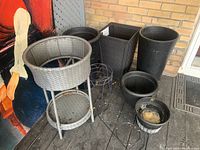 Multiple plastic planters in black color of different sizes, two black wire plant stands with circular design, and one gray woven wicker plant stand with two levels, all grouped together on a porch area.