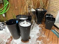Several black plastic planters on a wood deck in snowy conditions. Tall cylindrical planters are about 24 inches high, with two square pedestal style planters in the background.