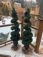 Two spiral spiral-shaped artificial topiary vases on an outdoor deck with light snow covering. Slightly worn resin vases with green artificial greenery and outdoor string lighting.