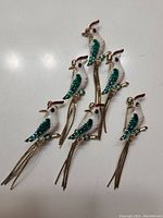Photo showing six bird brooches arranged on a white surface, highlighting design and details of the enamel and metal tassels.