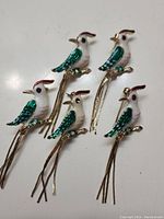 Five bird brooches with green, white and red enamel on metal. Each bird has long dangling metal tail feathers and detailed eyes and beak.