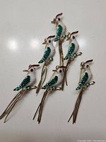 Six bird-shaped brooches with white and green enameled bodies and long gold-tone tail feathers, arranged on a white surface.