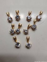 Ten faux diamond pendants with gold tone metal prong settings, displayed on a light surface; each pendant has a single clear round stone and a gold tone bail.