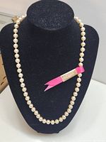 Full view of the 20-inch faux pearl necklace displayed on a black jewelry bust, showing the pearl strand and attached pink ribbon tag.