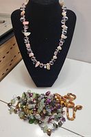 Photo showing one necklace displayed on black bust with pastel-colored pearl and gemstone beads, and other necklaces coiled on table below.