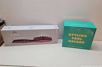 Sutra Ionic Heat Brush in original box next to PYT Styling Tool Holder box on a white surface against a beige wall.