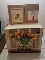 Photo showing three boxed items stacked: Blue Bird rain gauge, musical snowman figurine, and vase with tulips.