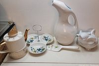 Photo showing five ceramic kitchenware items: white elephant teapot, white pitcher, divided floral dish, white and gold teapot, pig-shaped creamer on table