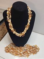 Three pearl necklaces displayed on a black velvet stand, showing creamy, golden-toned pearls and amber faceted beads in multiple strands.