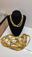 Photo shows a display stand with two yellow gemstone necklaces, one single strand and one multi-strand with silver metallic spacers laid out on a table.