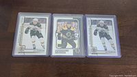 Three hockey cards in protective top loaders, two are Jake DeBrusk rookie cards in white Bruins uniform, one middle card is Boston Bruins player in black and yellow uniform.