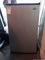 Closed front view of the silver and black Sanyo bar fridge with label on top left corner and Sanyo badge on upper right.