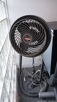 Front view of black Vornado floor fan next to a window and other items on floor.