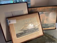 Photo showing two framed nautical prints of sailing ships on a glass table near a window with greenery outside.