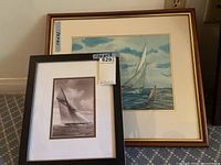 Both framed sailing-themed art pieces side by side on carpeted floor against wall, showing size difference; one larger colored print and one smaller black and white print.