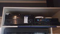 Shelf with Yamaha AV receiver RX-A720 with input and zone controls, and Sony media player device with blue lights and white remotes on top.