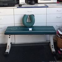 Green padded workout bench with a noticeable ripped area on the cushion and a U-shaped neck or arm support resting on top, situated indoors on a black rubber mat floor.