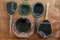 Five hand mirrors on wooden surface, showing variation in frame materials and styles including brass, guilloche, bakelite, and lucite.