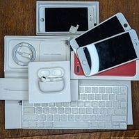 Photo showing Apple branded boxes and multiple iPhone display panels stacked on Apple Magic Keyboard with single AirPods Pro earbuds in charging case on top.