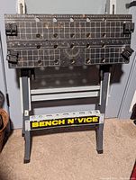Front view of folding work bench with composite wood top, integrated vice, steel frame, and BENCH N' VICE label.