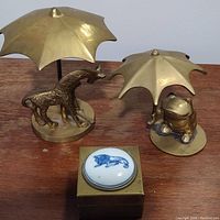 Photo showing brass giraffe and frog figurines each holding a brass umbrella, with a brass trinket box with porcelain lion plaque in front.