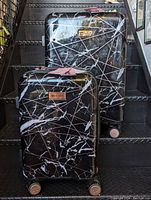 Front view of two Juicy Couture black hard shell rolling luggages with white marble-like pattern and gold lines, showing general overall design and size difference