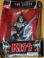 Front view of The Demon KISS figurine in original box with visible box damage and clear plastic window showing figure with bass guitar.